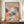 May include: Framed exhibition poster featuring abstract art with colorful polka dot patterns in coral, teal, navy and cream tones, displayed on wooden sideboard next to white ceramic vase and stacked books in cream-colored room with white molding