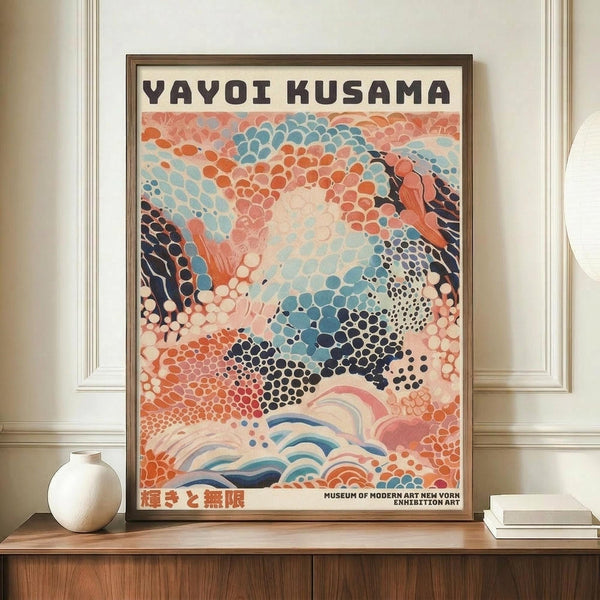May include: Framed exhibition poster featuring abstract art with colorful polka dot patterns in coral, teal, navy and cream tones, displayed on wooden sideboard next to white ceramic vase and stacked books in cream-colored room with white molding