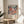 May include: Framed poster with colorful floral pattern in pink, coral, and blue tones displayed on round wooden side table next to beige upholstered chair. Poster features Japanese text reading "Yayoi Kusama" and "Japan 1987" below vibrant flower design. Natural lighting creates soft shadows on white wall. Green plant leaves visible in corner, creating cozy living room setting.
