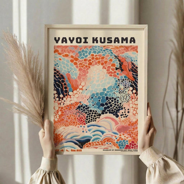 May include: Person holding framed poster featuring abstract art with colorful polka dot patterns in coral, teal, navy, and pink tones. Wave-like motifs flow across bottom. Museum of Modern Art New York text visible at bottom. Natural wood frame against white curtained background with dried pampas grass stems visible on left side.
