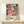 May include: Framed poster featuring colorful floral artwork in coral, pink, and teal tones with radiating flower patterns against navy background, displayed on light wood shelf next to small white wrapped gift with twine and greenery sprig, against cream wall