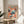 May include: Framed colorful abstract art poster with swirling patterns in orange, blue, and navy dots displayed on a round wooden side table next to a beige upholstered chair. Sunlight streams across white walls creating geometric shadows, with a green plant partially visible in the corner of this minimalist modern living space.