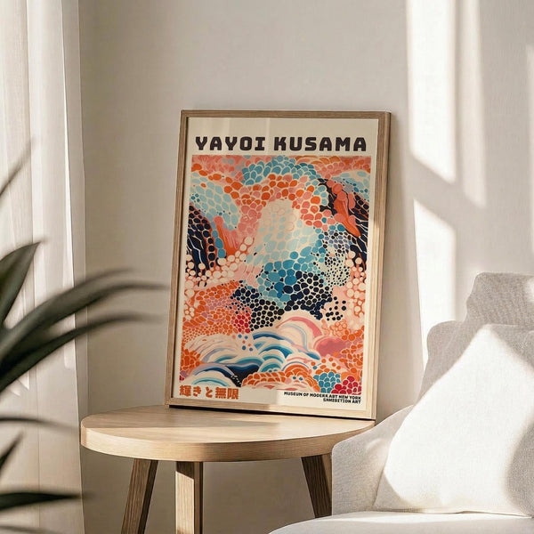 May include: Framed colorful abstract art poster with swirling patterns in orange, blue, and navy dots displayed on a round wooden side table next to a beige upholstered chair. Sunlight streams across white walls creating geometric shadows, with a green plant partially visible in the corner of this minimalist modern living space.