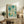 May include: Framed art print with abstract turquoise and teal bubble pattern leaning against white wall on rustic wooden sideboard. Print features scattered circles in various sizes with flowing watercolor effects and Japanese text below. Scene includes fern plant, stacked books, ceramic vessels, and beige upholstered chair with brown throw pillow. Natural lighting creates soft shadows on textured wall background.