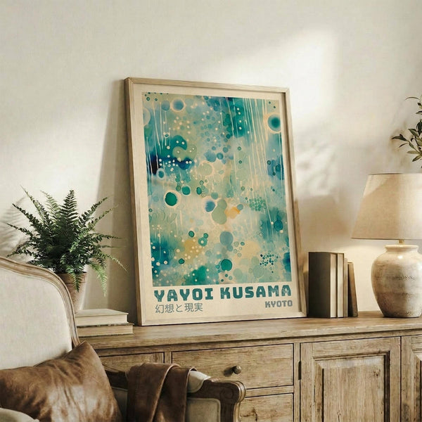 May include: Framed art print with abstract turquoise and teal bubble pattern leaning against white wall on rustic wooden sideboard. Print features scattered circles in various sizes with flowing watercolor effects and Japanese text below. Scene includes fern plant, stacked books, ceramic vessels, and beige upholstered chair with brown throw pillow. Natural lighting creates soft shadows on textured wall background.