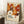 May include: Framed art print leaning against white wall next to wooden side table with white coffee cup. Poster features colorful retro floral pattern in orange, brown, and cream tones with geometric flower designs and polka dots. Text reads "Yayoi Kusama" with Japanese characters and "Musée d'Art Moderne de la Ville de Paris." Light wooden frame, cream matting, cozy room setting with textured rug and soft lighting.