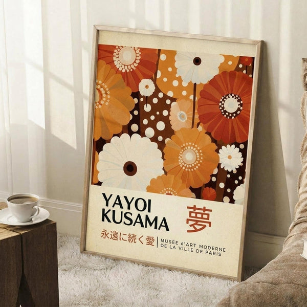 May include: Framed art print leaning against white wall next to wooden side table with white coffee cup. Poster features colorful retro floral pattern in orange, brown, and cream tones with geometric flower designs and polka dots. Text reads "Yayoi Kusama" with Japanese characters and "Musée d'Art Moderne de la Ville de Paris." Light wooden frame, cream matting, cozy room setting with textured rug and soft lighting.