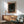 May include: Framed poster with dark cosmic design featuring swirling patterns, colorful dots, and celestial elements in orange, blue, and cream tones. Displayed above mid-century modern wood credenza with white lamp, next to sage green upholstered chair with wooden arms. Warm sunlight creates shadows on white wall in contemporary living room setting.