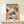May include: Framed art print leaning against white wall on light wood console table with small white gift box. Poster features abstract design with colorful dotted patterns in coral, blue, navy, and pink resembling organic cellular forms and flowing waves. Bold black text at top, natural wood frame, modern minimalist room setting.