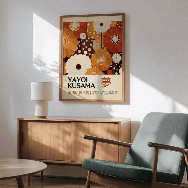 May include: Framed Yayoi Kusama exhibition poster with colorful polka-dotted flowers and circular patterns in orange, brown, and cream tones, displayed above a wooden sideboard with white lamp in modern living room setting with mint green armchair