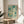 May include: Abstract art print with teal, blue and cream circular dots and dripping patterns in light wooden frame, displayed on round wooden side table next to beige upholstered chair. Sunlight streams across white wall creating soft shadows from nearby plant leaves. The artwork features organic bubble-like shapes in various sizes with flowing vertical elements throughout the composition.