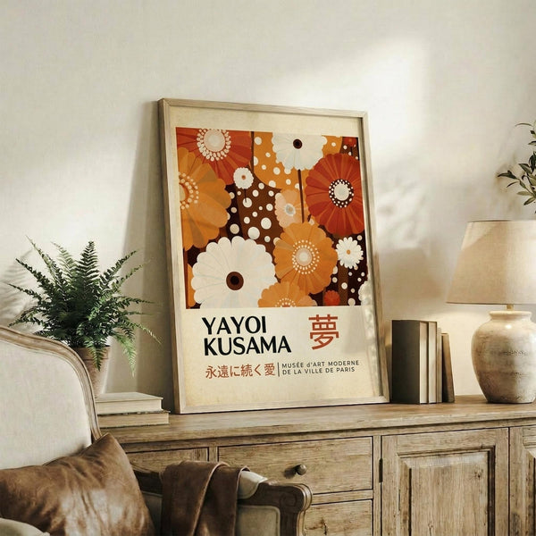 May include: Framed poster with colorful floral pattern in orange, brown and cream tones featuring polka dots and stylized flowers, displayed on rustic wooden sideboard next to fern plant, ceramic vase, and books in warm-toned living room setting with beige upholstered furniture.