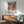 May include: Framed floral art print with colorful blooms in coral, pink, and teal against dark background, displayed above wooden sideboard with white ceramic lamp. Mid-century modern sage green armchair with wooden arms sits nearby, with sunlight casting striped shadows across white wall. Print features Japanese text and "JAPAN 1987" at bottom.