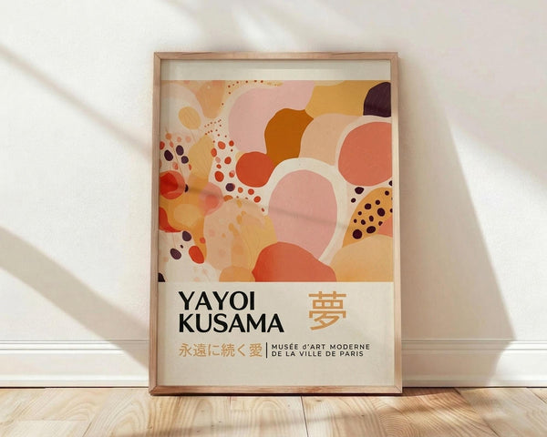 May include: Abstract art poster in wooden frame leaning against white wall on light wood floor. Artwork features organic flowing shapes in warm tones of coral, orange, pink, yellow and burgundy with scattered dots. Bottom text reads "YAYOI KUSAMA" with Japanese characters and "MUSÉE D'ART MODERNE DE LA VILLE DE PARIS". Soft natural lighting creates diagonal shadows across textured white wall.