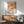 May include: Framed abstract art poster with warm orange, pink, and coral circular shapes and polka dot patterns, displayed above a wooden sideboard with ceramic lamp. Mid-century modern living room setting features sage green upholstered chair with wooden arms, round coffee table, and natural sunlight creating geometric shadows on white walls.