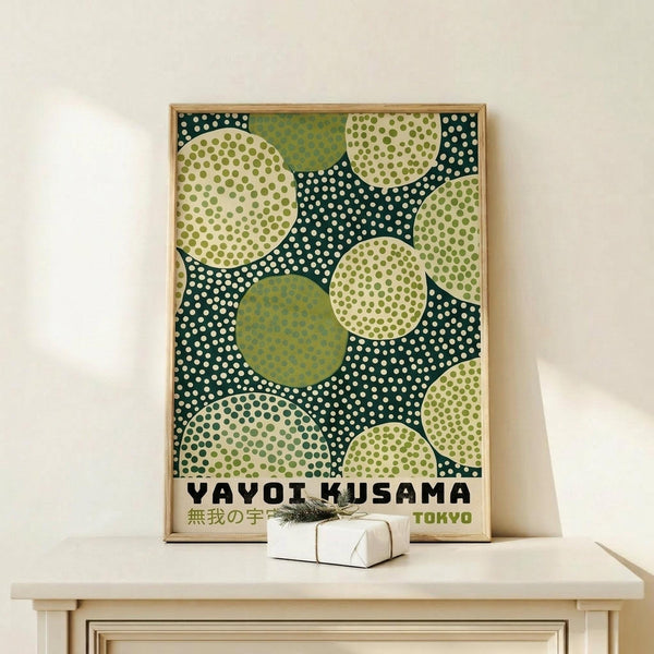 May include: Framed art print with green polka dot pattern featuring overlapping circles in various shades of green and cream on dark teal background, displayed on light wood console table next to small wrapped gift with sprig decoration against cream wall