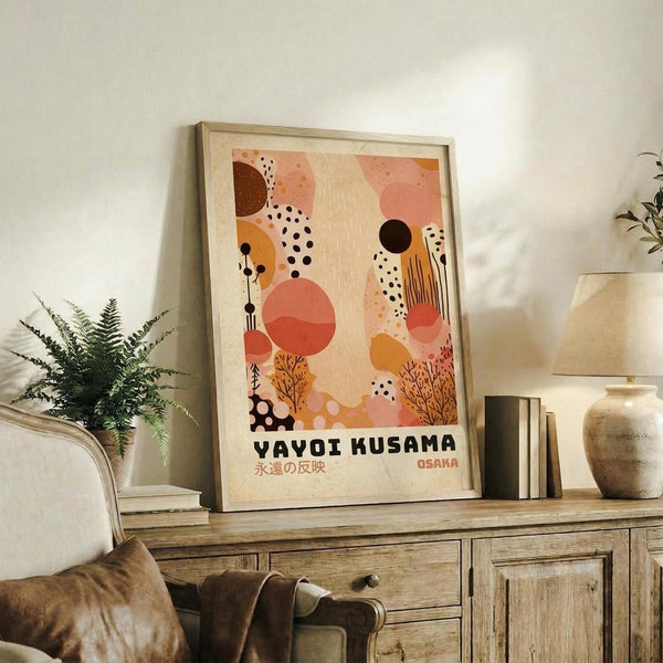 May include: Framed abstract art poster with organic shapes in coral, orange, brown, and cream colors featuring dots, circles, and botanical elements, displayed on rustic wooden sideboard next to fern plant, ceramic vase, and stacked books in warm-toned living room setting.