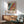 May include: Colorful abstract art poster in wooden frame hanging above modern credenza with table lamp. Green mid-century chair sits nearby. Poster features flowing patterns in black, blue, yellow, pink and orange with polka dots. Japanese text and museum information below artwork. Bright natural light creates shadows across white wall and wooden furniture.