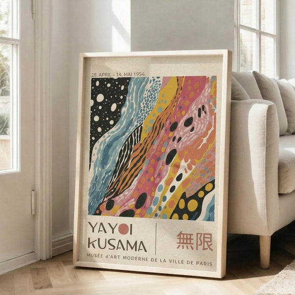 May include: A framed exhibition poster featuring Yayoi Kusama's abstract artwork with flowing organic shapes in vibrant colors - black starry section, blue waves, pink and orange swirls with polka dots. Text shows dates "28. APRIL - 14. MAI 1954" and "YAYOI KUSAMA" with "MUSÉE D'ART MODERNE DE LA VILLE DE PARIS" below. The light wood frame leans against white walls near large windows, positioned next to a beige sofa on hardwood floors in a bright, minimalist living roo