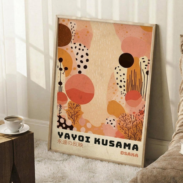 May include: Framed abstract art print featuring warm coral, orange, and cream organic shapes with black dots, botanical elements, and Japanese text reading "Yayoi Kusama" and "Osaka." Print leans against white wall next to wooden side table with coffee cup and cozy blanket in softly lit room setting.