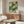May include: Modern living room mockup showing framed Yayoi Kusama-inspired poster with green and cream polka dot circles on dark teal background, displayed above mid-century walnut credenza with sliding doors. Scene includes white curved chair, brass floor lamp, white table lamp, and oval glass coffee table on wooden base against neutral beige walls.