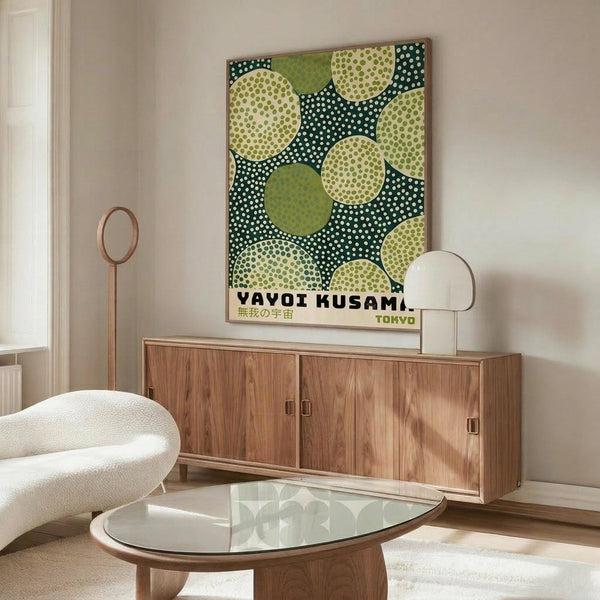 May include: Modern living room mockup showing framed Yayoi Kusama-inspired poster with green and cream polka dot circles on dark teal background, displayed above mid-century walnut credenza with sliding doors. Scene includes white curved chair, brass floor lamp, white table lamp, and oval glass coffee table on wooden base against neutral beige walls.