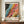 May include: Framed vintage-style exhibition poster featuring abstract flowing patterns in vibrant colors - black starry section transitioning to blue, orange, pink and yellow organic shapes with dots and swirls. Text shows "YAYOI KUSAMA" and Japanese characters below colorful artwork. Poster displayed on wooden sideboard beside white ceramic vase and stacked books in bright room with white walls and molding.