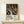 May include: Framed art print on light wood surface featuring abstract flowing lines and colorful dots on black background, with "YAYOI KUSAMA" text and Japanese characters below. Small white wrapped gift box sits nearby. Poster shows organic curved forms in tan and rainbow colors dotted throughout dark space, creating cosmic or cellular appearance. Natural lighting on neutral wall background.