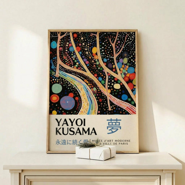 May include: Framed art print on light wood surface featuring abstract flowing lines and colorful dots on black background, with "YAYOI KUSAMA" text and Japanese characters below. Small white wrapped gift box sits nearby. Poster shows organic curved forms in tan and rainbow colors dotted throughout dark space, creating cosmic or cellular appearance. Natural lighting on neutral wall background.
