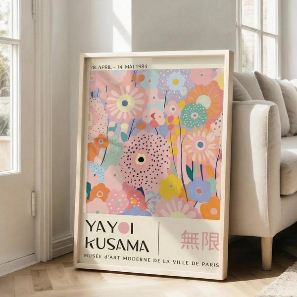 May include: Framed exhibition poster featuring colorful abstract floral design with pastel pink, blue, yellow and orange flowers and polka dot patterns, displayed against white wall next to beige sofa in bright living room with large windows and natural lighting