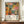 May include: Framed exhibition poster featuring vibrant botanical artwork with colorful abstract flowers, leaves, and organic shapes in pink, orange, yellow, and green tones. Text reads "YAYOI KUSAMA" and "JAPAN 1987" with Japanese characters. Poster displayed on wooden sideboard next to white ceramic vase and stacked books in elegant room with cream walls and white trim.