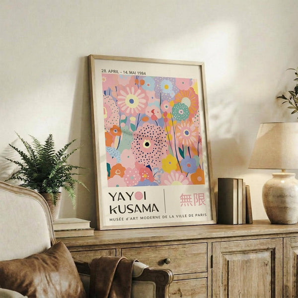 May include: Framed colorful floral art print displayed on rustic wooden sideboard next to fern plant, books, and ceramic vases. The poster features vibrant pink, blue, orange and yellow abstract flowers with polka dot patterns against pastel background. Natural lighting creates soft shadows on white wall. Exhibition-style artwork with text beneath botanical design.
