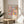 May include: Colorful art poster in wooden frame displayed on round wooden side table next to beige upholstered chair. Artwork features vibrant floral and dotted patterns in pink, blue, yellow, and orange with exhibition text below. Soft natural lighting creates shadows on white wall. Green plant leaves visible in corner.