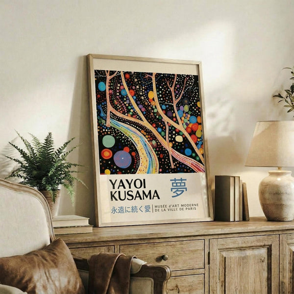 May include: Framed poster with colorful abstract art featuring flowing tree-like forms with multicolored dots and circles on black background, displaying "YAYOI KUSAMA" text below artwork. Light wood frame sits on rustic wooden sideboard alongside fern plant, books, and ceramic vase in bright modern living room setting with beige furniture and white walls.