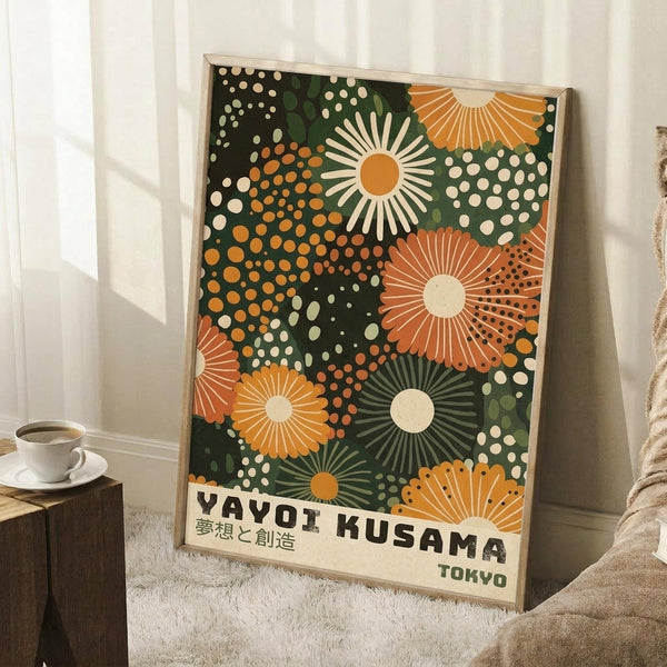 May include: Framed poster with vibrant abstract floral pattern in orange, green, and cream colors on dark background, featuring radiating sunburst designs and polka dots. Shows Japanese text at bottom. Displayed leaning against white wall corner next to coffee cup on wooden surface and textured throw blanket in bright, naturally-lit room setting.