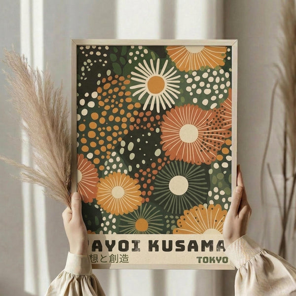 May include: Person holding framed art print with abstract floral sunburst pattern in orange, green, and cream colors on dark background. Japanese text at bottom reads "AYOI KUSAMA" and "TOKYO". Light wood frame, neutral beige clothing, dried pampas grass visible, soft natural lighting against white curtained window.