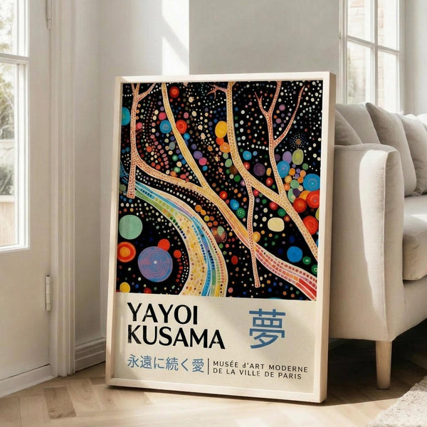 May include: Framed art poster featuring Yayoi Kusama's distinctive polka dot style with black background, colorful flowing ribbons, circles and dots in vibrant blues, reds, yellows and greens. White frame leaning against light gray wall next to beige sofa in bright room with large windows. Japanese text and museum attribution visible at bottom of poster.