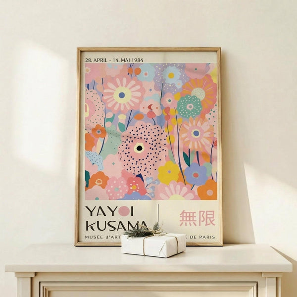 May include: Framed colorful floral art print leaning against white wall on light wood surface. Artwork features pastel pink, blue, orange and green abstract flowers with polka dots. Small white gift box with twine sits in front of frame. Print shows exhibition text reading "YAYOI KUSAMA" and "MUSEE d'ART DE PARIS" with dates "28. APRIL - 14. MAI 1984". Light wood frame complements minimal neutral decor setting.