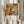 May include: Person holding framed vintage Japanese art poster featuring vibrant abstract floral design with orange, pink, and teal circular blooms, dotted patterns, and botanical elements against dark background. Light wood frame, cream matting with black text including "YAYOI KUSAMA" and "JAPAN 1987" with Japanese characters. Dried wheat stems visible to left, neutral curtained background, person wearing beige ruffled sleeve top.