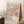 May include: Colorful abstract floral poster in light wood frame leaning against white wall corner. Artwork features pink, blue, yellow, and orange flowers with dotted patterns and polka dots in playful, modern style. Text shows dates "28. APRIL - 14. MAI 1984" and "YAYOI KUSAMA MUSÉE D'ART MODERNE DE LA VILLE DE PARIS" with Japanese characters. White coffee cup on wooden block and cream knit blanket visible nearby on fluffy white rug.