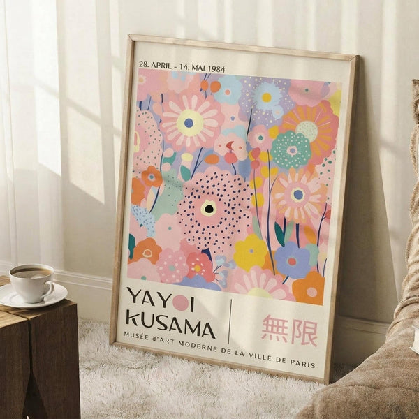 May include: Colorful abstract floral poster in light wood frame leaning against white wall corner. Artwork features pink, blue, yellow, and orange flowers with dotted patterns and polka dots in playful, modern style. Text shows dates "28. APRIL - 14. MAI 1984" and "YAYOI KUSAMA MUSÉE D'ART MODERNE DE LA VILLE DE PARIS" with Japanese characters. White coffee cup on wooden block and cream knit blanket visible nearby on fluffy white rug.