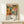 May include: Framed poster with vibrant botanical illustration featuring colorful abstract flowers, mushrooms, and organic shapes in orange, pink, green, and yellow tones. Text reads "YAYOI KUSAMA JAPAN 1987" with Japanese characters. Displayed on light wood console with small wrapped gift box. Natural wood frame against cream wall in minimalist room setting.