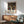 May include: Yayoi Kusama exhibition poster in thin wood frame hanging on white wall above mid-century modern wooden credenza with white ceramic lamp. Features black background with colorful flowing ribbons, polka dots, and circular shapes in vibrant blues, oranges, yellows, and greens. Japanese text below artwork. Sage green upholstered chair with wooden arms partially visible, natural sunlight creating shadows across the minimalist room setting.