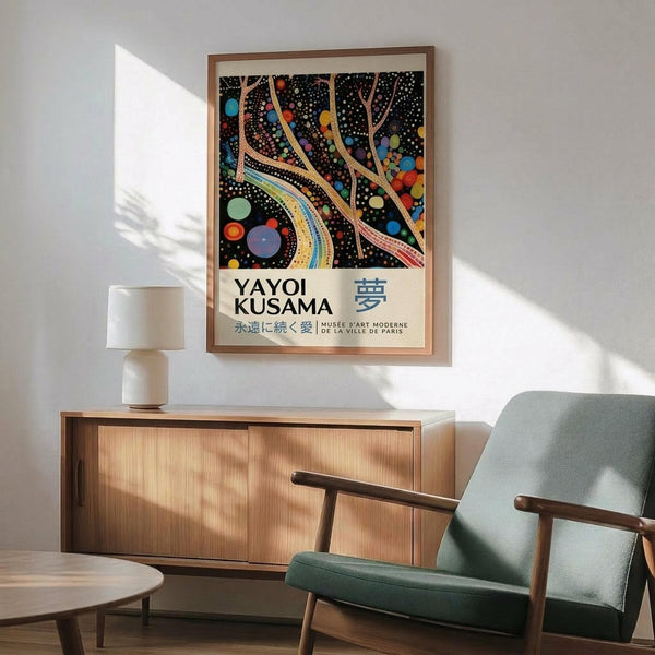 May include: Yayoi Kusama exhibition poster in thin wood frame hanging on white wall above mid-century modern wooden credenza with white ceramic lamp. Features black background with colorful flowing ribbons, polka dots, and circular shapes in vibrant blues, oranges, yellows, and greens. Japanese text below artwork. Sage green upholstered chair with wooden arms partially visible, natural sunlight creating shadows across the minimalist room setting.