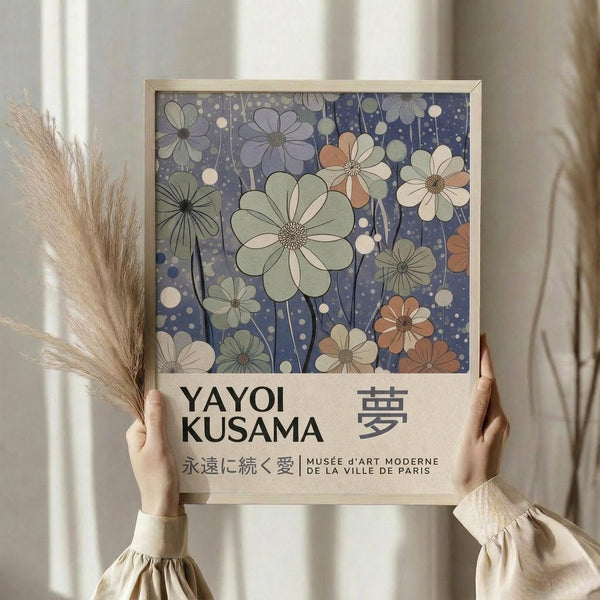 May include: Two hands holding a framed art print featuring stylized flowers in muted green, coral, and cream tones against a deep blue background with polka dots. The print shows Japanese text and museum attribution below the floral design. A bundle of dried pampas grass sits nearby against flowing white curtains in soft natural lighting.