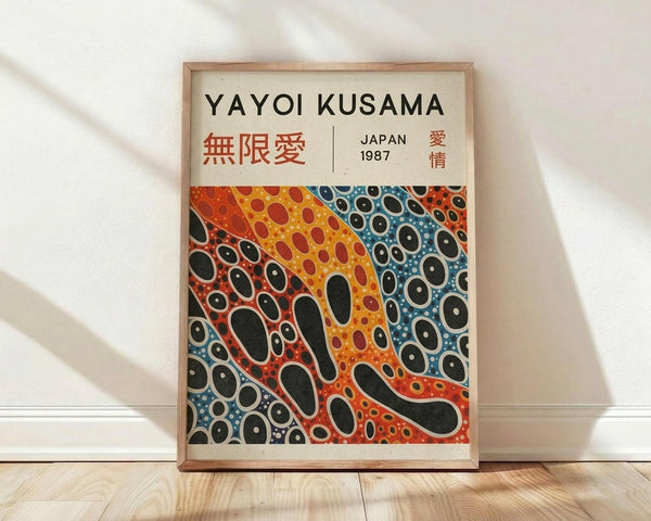 May include: Framed poster print leaning against white wall on light wooden floor. The artwork features Yayoi Kusama's signature polka dot style in vibrant red, orange, yellow, and blue colors with flowing organic shapes and circular patterns. Japanese text appears at top along with "JAPAN 1987". Natural wooden frame surrounds the cream-colored poster. Soft natural lighting creates gentle shadows on the white textured wall.
