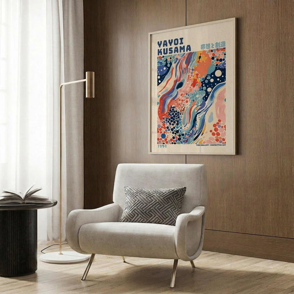 May include: Colorful abstract art poster with flowing waves, dots, and organic shapes in blues, oranges, and pinks, displayed in light wood frame on wood paneled wall above beige upholstered armchair with geometric patterned pillow, brass floor lamp, and dark round side table with open book in modern neutral-toned room.
