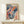 May include: Framed colorful abstract art poster featuring flowing waves, polka dots, and organic shapes in navy blue, coral red, teal, and pink tones with "YAYOI KUSAMA" text and Japanese characters at top. Light wood frame sits on pale wooden console table against white wall, with small white wrapped gift box placed in front.