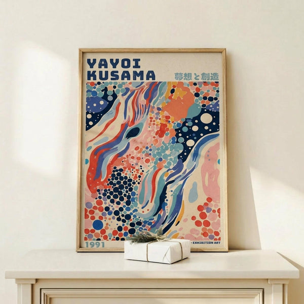 May include: Framed colorful abstract art poster featuring flowing waves, polka dots, and organic shapes in navy blue, coral red, teal, and pink tones with "YAYOI KUSAMA" text and Japanese characters at top. Light wood frame sits on pale wooden console table against white wall, with small white wrapped gift box placed in front.