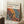 May include: Framed art print leaning against white wall corner on textured cream rug. Poster features Yayoi Kusama's name in black text with Japanese characters, dated Japan 1987. Artwork shows organic flowing shapes in vibrant orange, yellow, red and blue with black polka dots in Kusama's signature style. Light wood frame, cozy room setting with coffee cup on wooden block nearby and beige textiles.